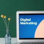A laptop displaying 'Digital Marketing' with a minimalist floral decor in a glass vase.